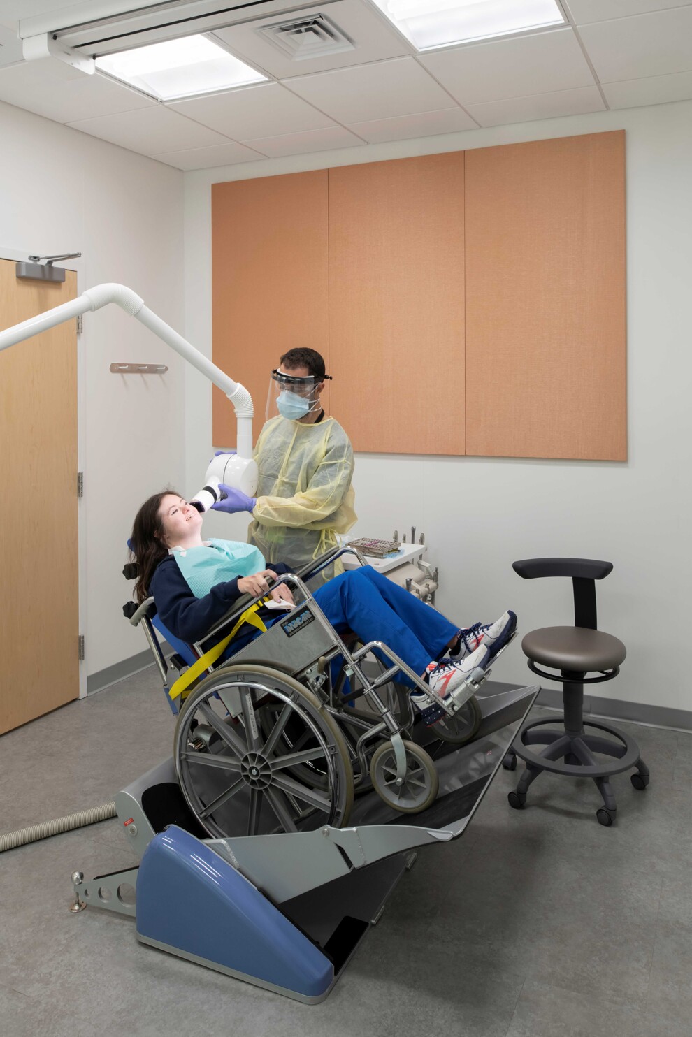 A patient using a wheelchair receives dental treatment in a wheelchair recliner.